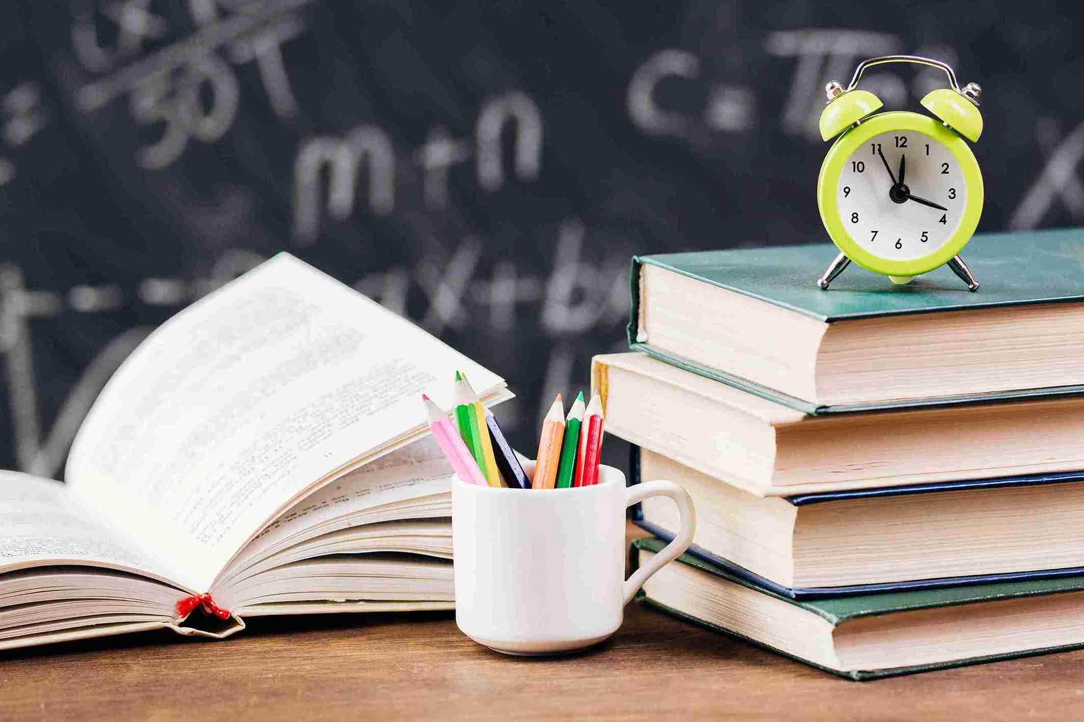 Open textbook, a cup of colored pencils, stacked books, and an alarm clock on a desk in front of a chalkboard.